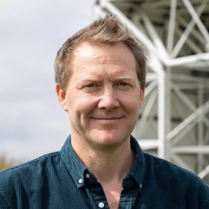 A man with short, light brown hair and a blue corduroy shirt smiles at the camera outdoors, with a blurred metal structure and sky in the background.