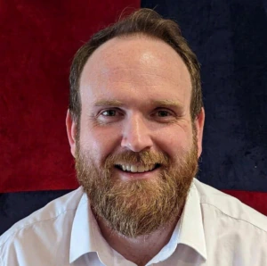 A bearded man with light brown hair wearing a white shirt smiles at the camera. The background features a red and dark blue color block pattern.