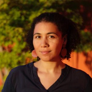 A woman with curly hair, wearing a dark blue top and large round earrings, stands outdoors in warm sunlight. Green foliage and an orange wall are blurred in the background.