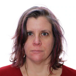 A woman with shoulder-length brown hair with purple highlights, wearing a red top and gold necklace, stares directly at the camera against a plain white background.