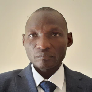 A man in a dark blue suit, white shirt, and blue tie stands against a plain, light-colored background, looking directly at the camera with a neutral expression.