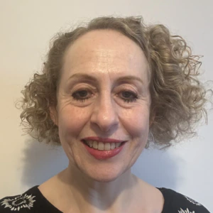 A woman with short, curly blonde hair and red lipstick smiles at the camera. She is wearing a black top with white flower patterns and is standing in front of a plain light-colored wall.