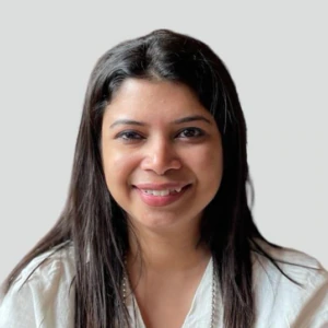 A woman with straight dark hair, wearing a white blouse, is smiling and facing the camera against a plain light gray background.