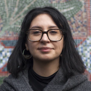 A woman with shoulder-length black hair and glasses stands in front of a colorful mosaic wall. She is wearing a gray sleeveless sweater over a black top and is looking at the camera with a slight smile.