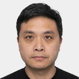 Portrait of an adult man with short black hair, wearing a plain black shirt, looking directly at the camera with a neutral expression against a white background.