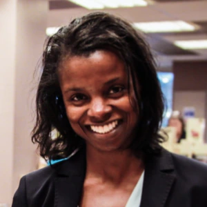 A woman with shoulder-length dark hair, wearing a black blazer and light blue blouse, stands indoors smiling. Books and shelves are visible in the blurred background, suggesting a bookstore or library setting.