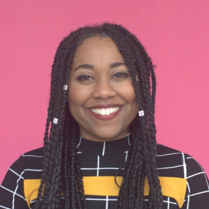 A smiling woman with long braided hair and bead accents wears a black and yellow checkered top, standing in front of a solid pink background.