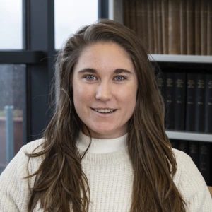 A woman with long brown hair wearing a cream-colored sweater smiles at the camera. She is indoors, in front of a window and shelves filled with books.