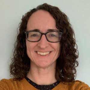 A person with curly brown hair and glasses smiles at the camera. They are wearing a mustard yellow sweater over a black shirt with small white designs, standing against a plain light-colored background.