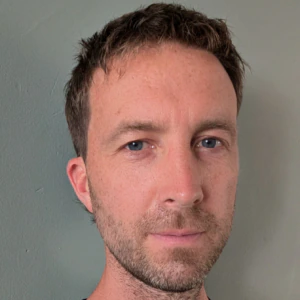 A man with short brown hair and light beard wearing a black crew neck shirt stands in front of a plain light gray wall, looking directly at the camera with a neutral expression.