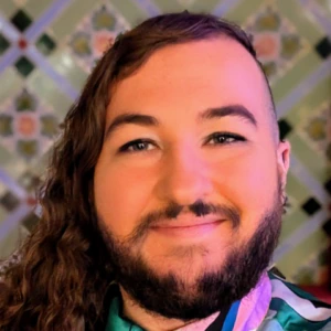A person with long, curly brown hair on one side and a beard smiles at the camera. They wear a colorful, patterned shirt and decorative pins, with a floral tile background behind them.