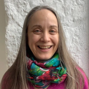 A woman with long gray hair smiles at the camera. She is wearing a bright pink top and a colorful scarf with green, red, and blue patterns. The background is a textured white wall.