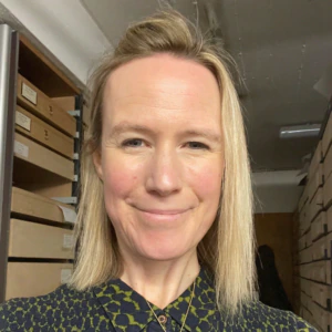 A woman with straight blond hair and a patterned dark green and blue top smiles in a room with shelves of labeled archive boxes. She wears a gold necklace with a round pendant.