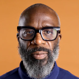 A middle-aged man with a gray beard and black glasses wears a navy blue collared shirt, standing against an orange background.