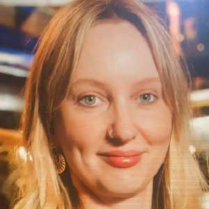 A woman with long blonde hair and blue eyes smiles softly. She is wearing a floral top and geometric earrings, with a nose ring. The background has warm lights and a blurred indoor setting.