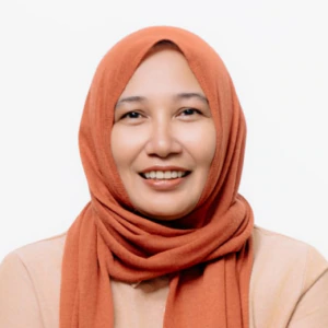 A woman wearing a peach-colored top, olive green pants, and a terracotta hijab sits on a stool, smiling, with her hands crossed over her knee against a plain white background.