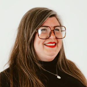A woman with long brown hair, wearing large square glasses, red lipstick, a black turtleneck, and a silver pendant necklace, smiles while looking slightly to the side against a plain white background.