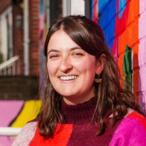 A woman with long brown hair smiles while standing against a brightly painted brick wall featuring colorful, abstract designs. She wears a red, pink, and white striped sweater and gold earrings.