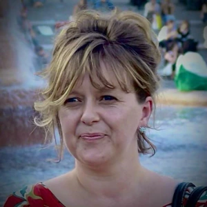 A woman with wavy, light brown hair and bangs smiles outdoors in front of a fountain. She is wearing a patterned top and earrings, with blurred people and water in the background.