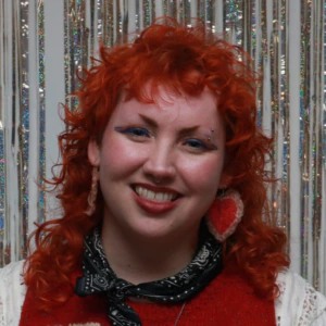 A person with curly red hair, blue eye makeup, and heart-shaped earrings smiles in front of shimmering silver streamers.