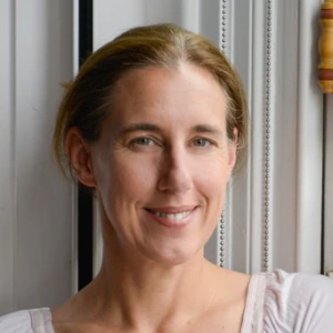 A woman with light brown hair pulled back, wearing a light pink long-sleeve top, stands indoors by a window with her arms crossed, smiling at the camera.