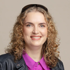 Smiling person with curly blonde hair wearing a black leather jacket, a bright purple shirt, and sunglasses on their head, posed against a plain beige background.