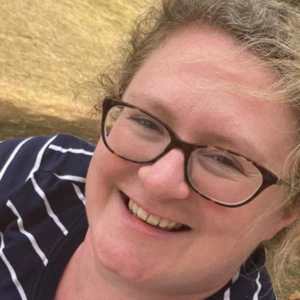 A person with curly blond hair and glasses smiles outdoors, wearing a navy blue and white striped shirt. Grass and trees are visible in the background.