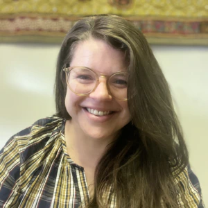 A person with long brown hair and glasses smiles while wearing a plaid shirt, sitting in front of a white wall and a colorful, patterned tapestry.