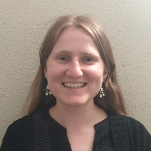 A person with long light brown hair is smiling at the camera, wearing a black textured top, a black cardigan, and dangly earrings, standing in front of a plain beige wall.