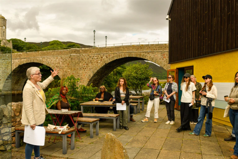 A group of people listens to a woman speaking outdoors near stone picnic tables, with a stone bridge and building in the background. Some hold papers. The sky is cloudy.