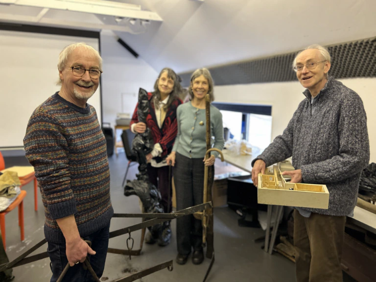 Four older adults stand in a well-lit room, smiling at the camera. One holds metal tools, another has a box with compartments, and two women at the back carry bags. The setting appears informal and creative.