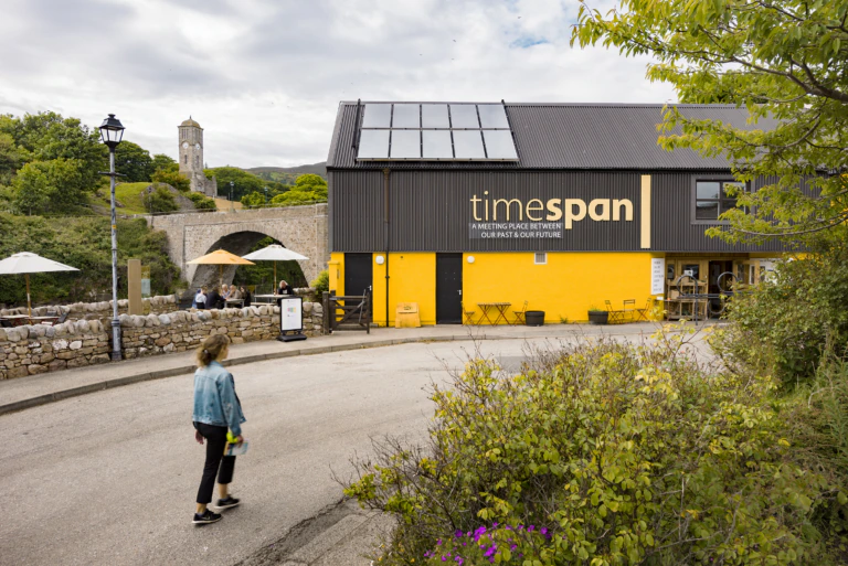 A person walks on a curved road past a yellow and black building labeled Timespan, with outdoor seating, trees, a stone bridge, and a church tower visible in the background under a cloudy sky.