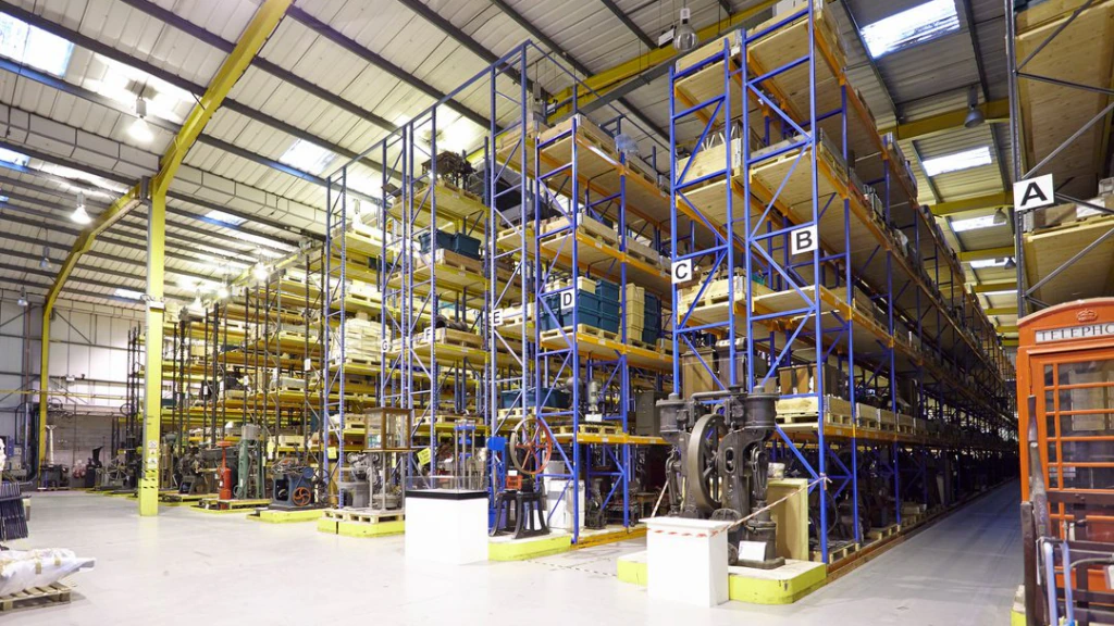 Large warehouse with tall blue and yellow storage racks filled with various items, labeled A to D. A person in a safety vest walks in the aisle. The space is brightly lit and organized.