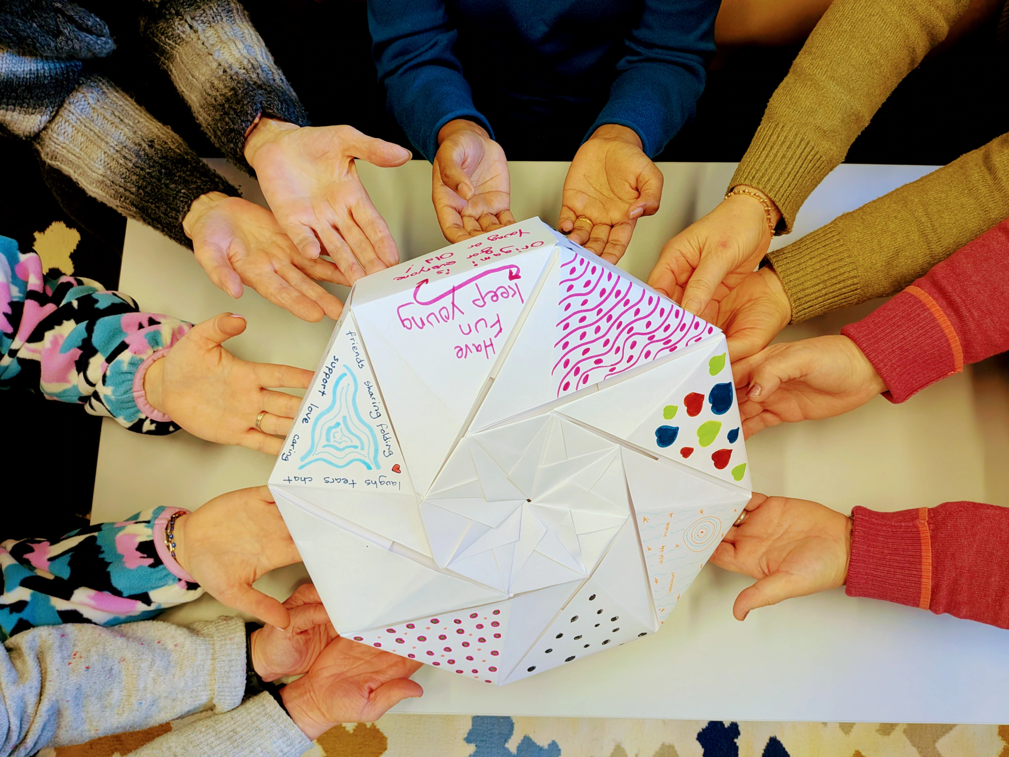 Eight people hold out their hands around a large, decorated paper fortune teller with colorful patterns and handwritten messages on a white table.