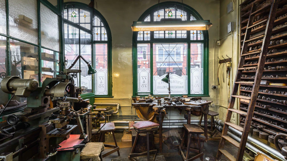 A vintage workshop with wooden stools, metal tools, machinery, and wooden shelves, set in a room with arched windows and stained glass, letting in daylight. The space looks organized but heavily used.