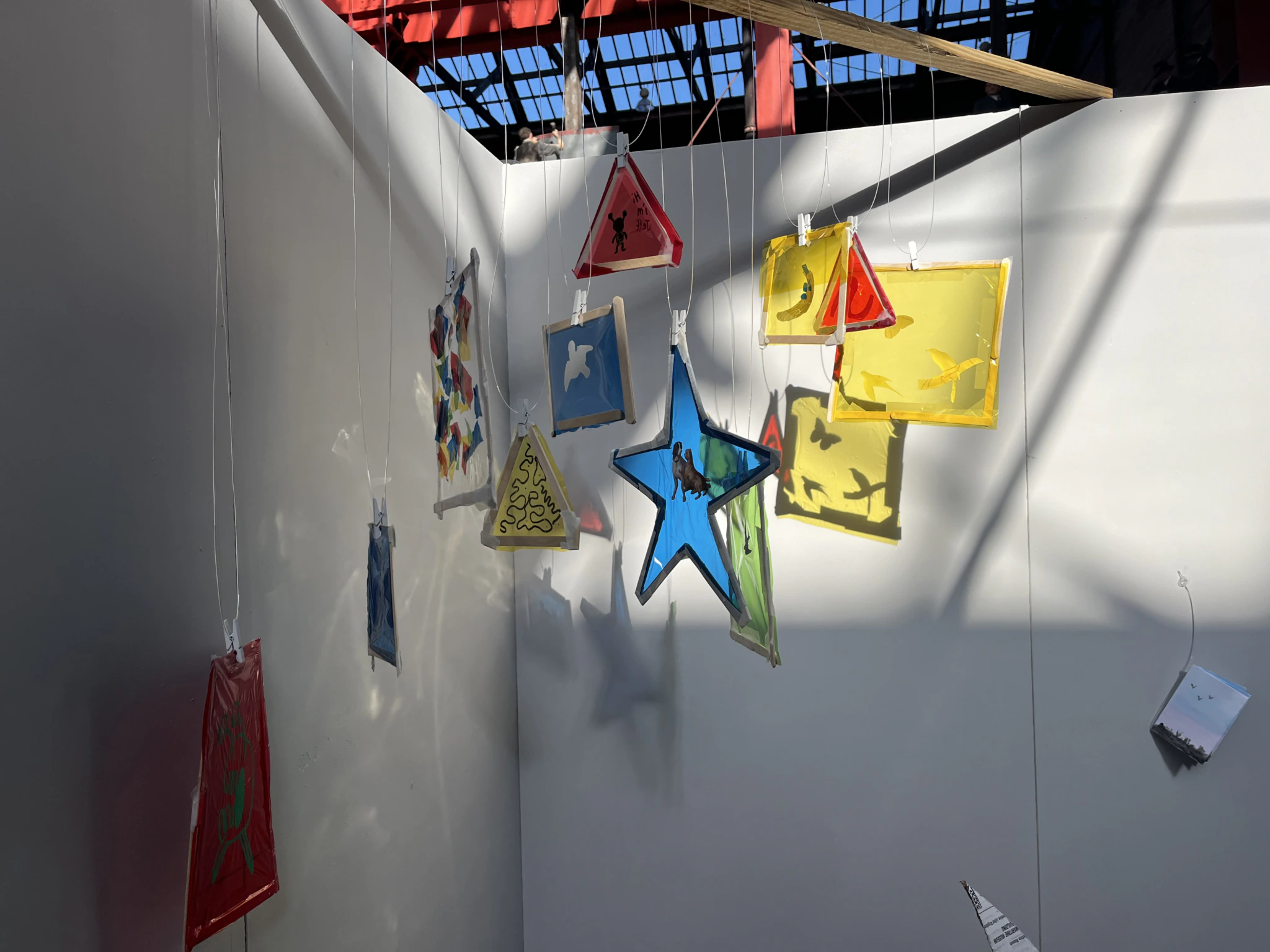 Colorful translucent shapes, including stars, triangles, and rectangles, hang from strings in a sunlit corner with white walls and a partially open roof casting shadows.