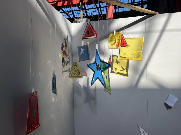 Colorful translucent shapes, including stars, triangles, and rectangles, hang from strings in a sunlit corner with white walls and a partially open roof casting shadows.