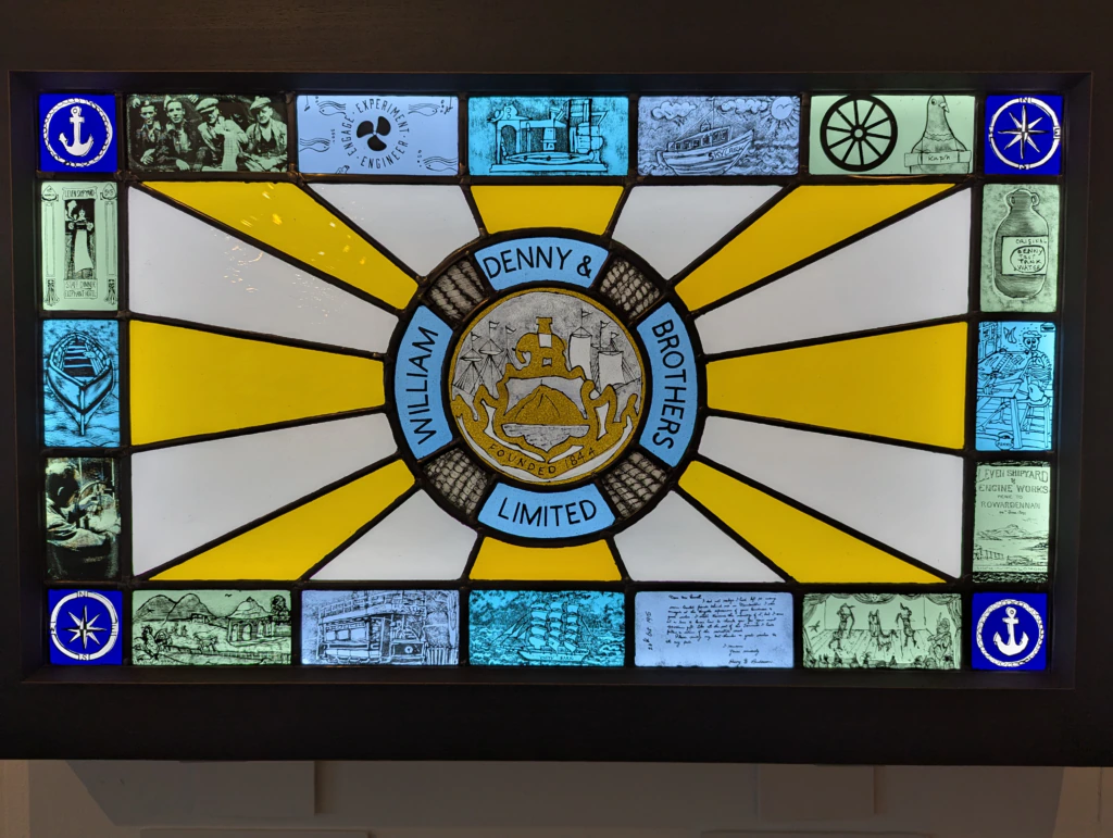 Stained glass window with the William Denny & Brothers Limited crest in the center, surrounded by nautical-themed illustrations and symbols, including anchors and ships. Yellow and white rays radiate from the center.