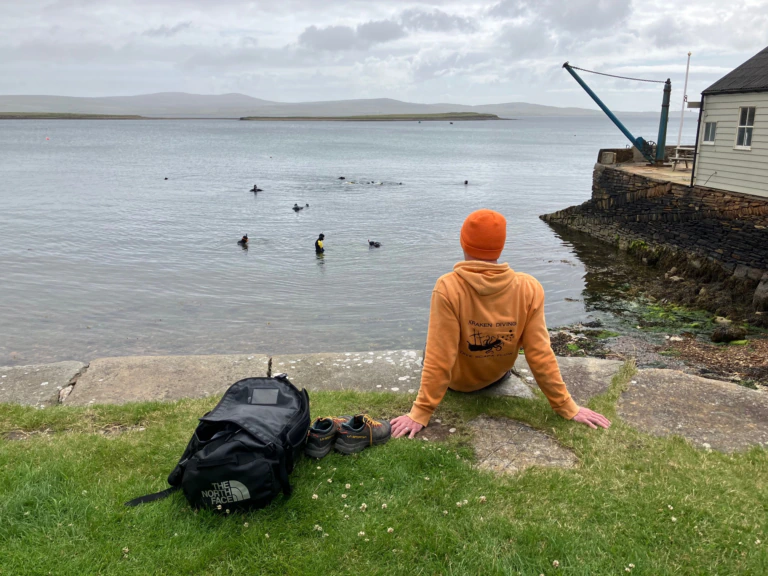 A person in an orange hoodie and hat sits by the water, facing swimming ducks. A black backpack and shoes are beside them on the grass. The sky is cloudy, and a stone wall lines the shore.