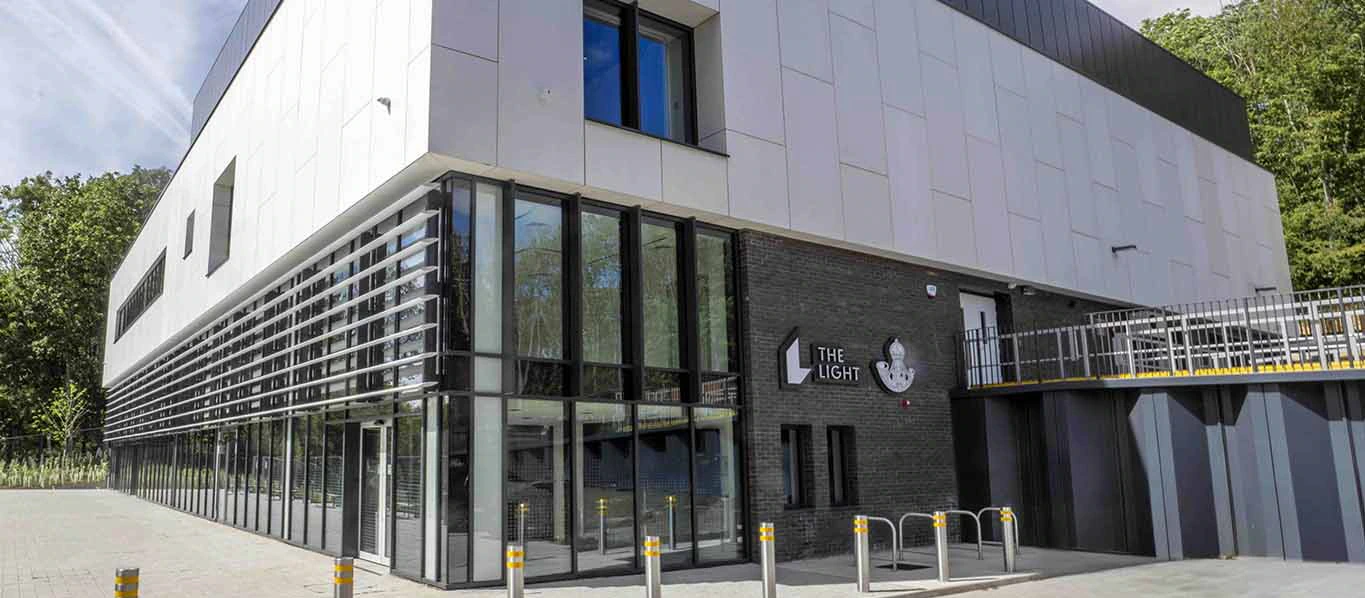 A modern building with large windows and a white facade under a blue sky; signs read The Light and The University of Huddersfield. Bollards stand in front of the entrance.