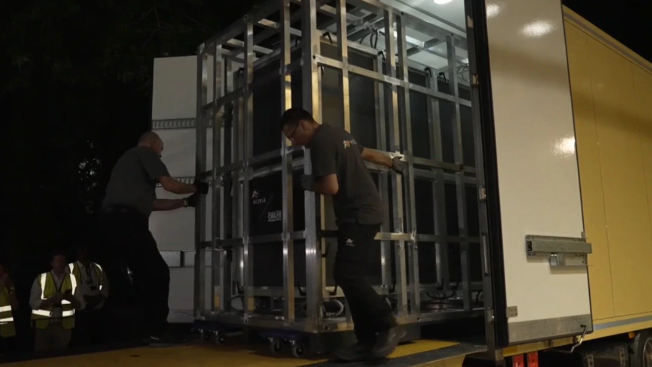 Two workers are unloading a large, metal-framed container from a yellow truck at night. The area is dimly lit, and several people in reflective vests are visible in the background.