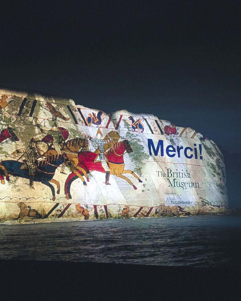 A nighttime projection of the Bayeux Tapestry with Merci! and The British Museum displayed on a large outdoor wall by the water.