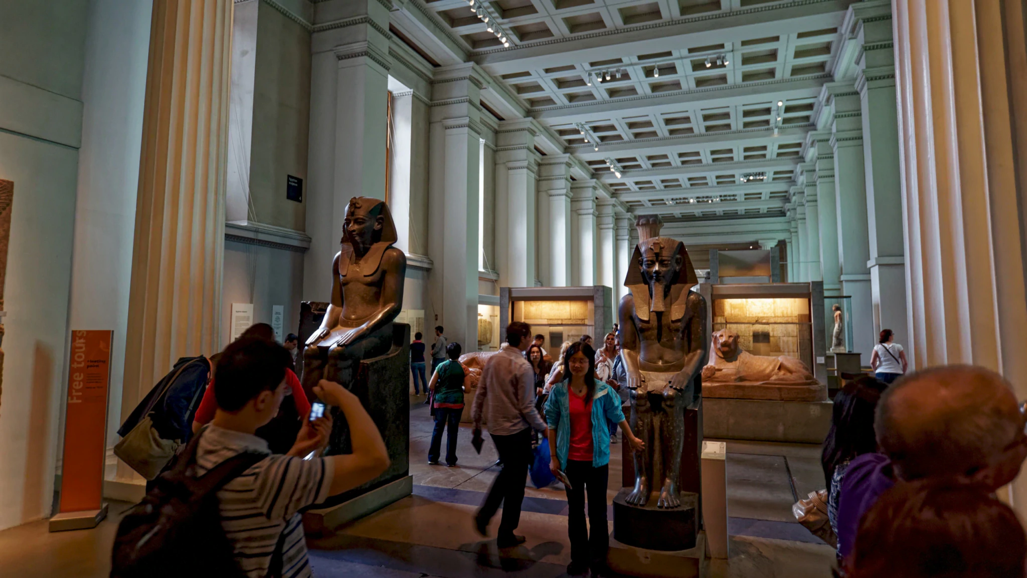 Visitors walk and take photos among large ancient Egyptian statues in a spacious museum hall with tall columns and a high, coffered ceiling.