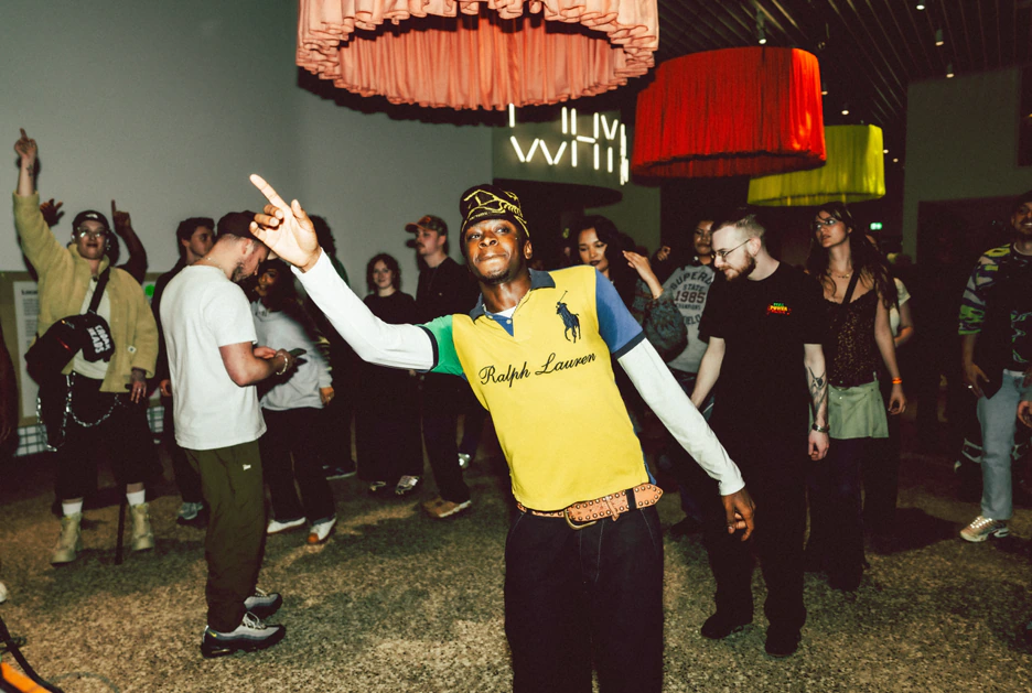 A person in a yellow Ralph Lauren shirt points upward while dancing at a crowded indoor event; others in the background are mingling, dancing, and enjoying the atmosphere under colorful hanging lights.