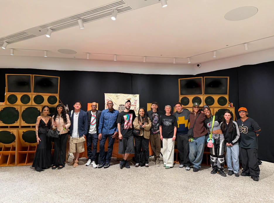 A group of people stand in a row, smiling in front of large speaker stacks and a banner in a brightly lit indoor space.