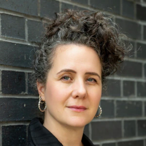 A person with curly dark hair in a bun, wearing hoop earrings and a black collared shirt, stands against a dark brick wall, looking confidently at the camera.
