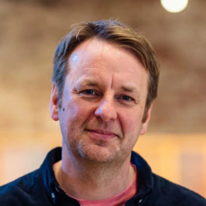 A middle-aged man with short brown hair and a slight smile, wearing a dark shirt over a pink t-shirt, stands indoors with a blurred background.