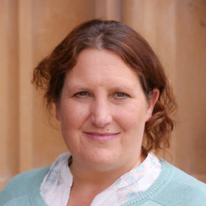 A woman with wavy brown hair smiles gently at the camera. She is wearing a light blue sweater over a white patterned blouse. The background is soft and beige, possibly a wall or building.