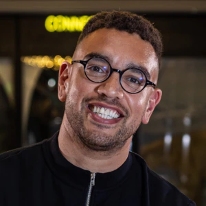 A smiling man with short curly hair, round glasses, and a trimmed beard wearing a black zip-up top stands indoors with blurred lights in the background.