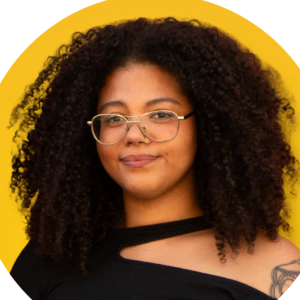 A person with curly hair, glasses, and a nose ring smiles slightly. They wear a black top with a cutout shoulder and a visible tattoo. The background is a vibrant yellow circle.